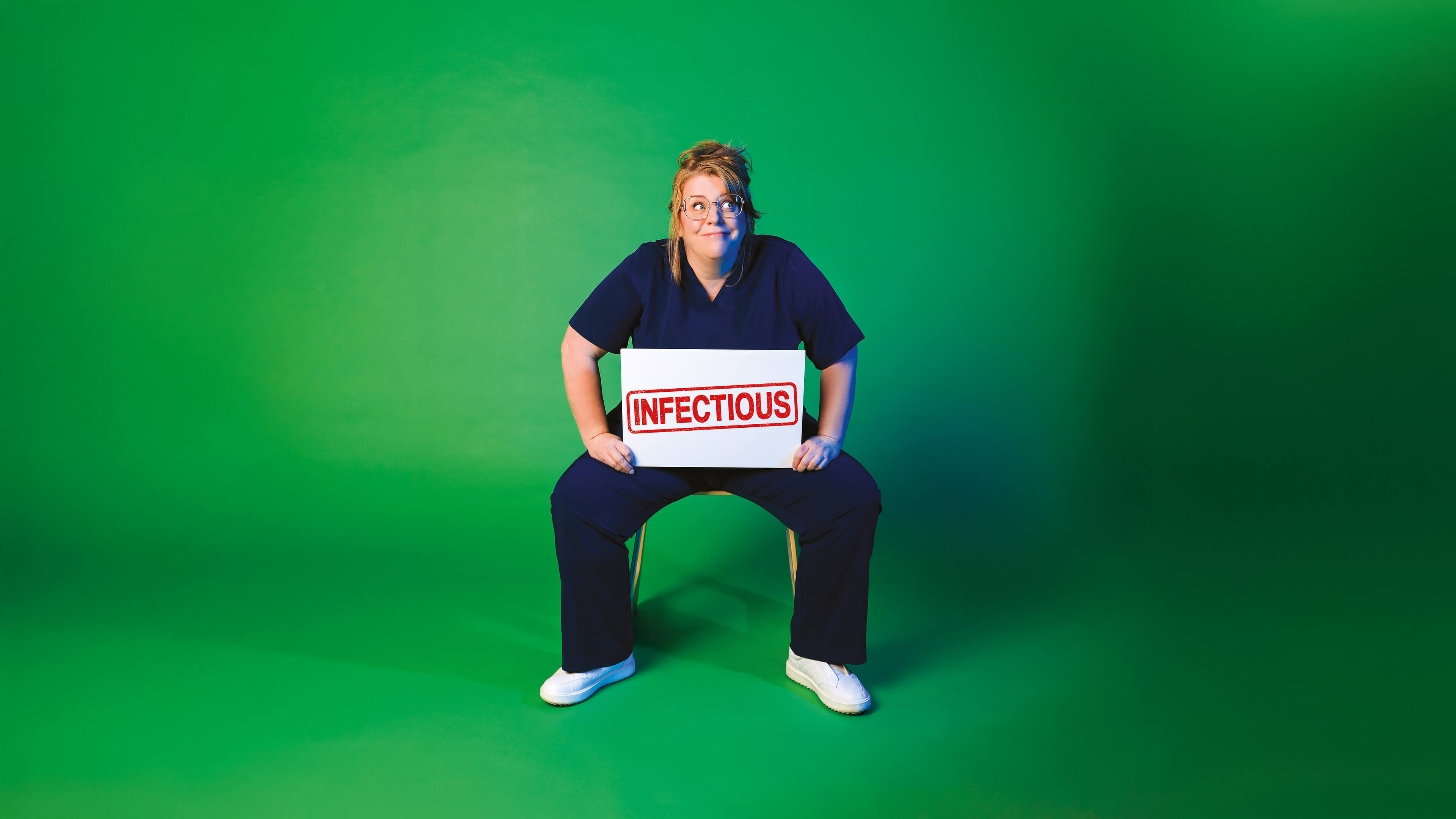 Person in navy medical scrubs sitting on a chair against a green background, holding a white sign with the word ‘INFECTIOUS’ stamped in red.