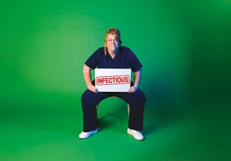 Person in navy medical scrubs sitting on a chair against a green background, holding a white sign with the word ‘INFECTIOUS’ stamped in red.