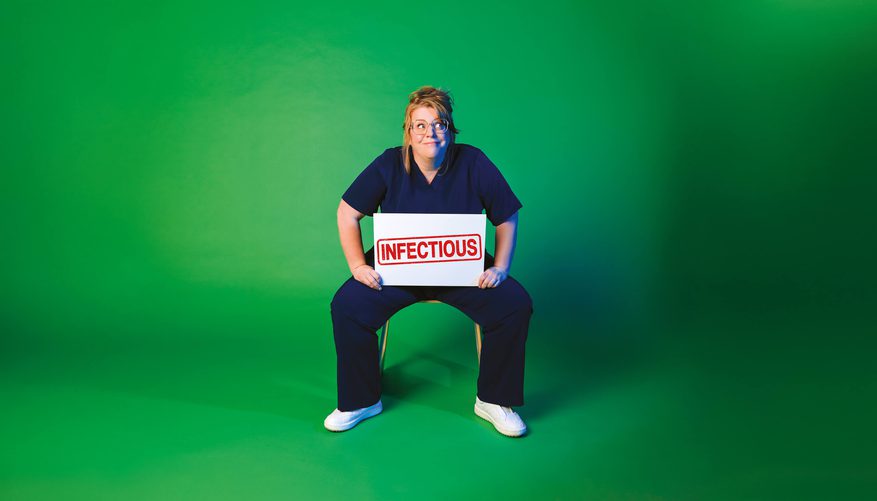 Person in navy medical scrubs sitting on a chair against a green background, holding a white sign with the word ‘INFECTIOUS’ stamped in red.