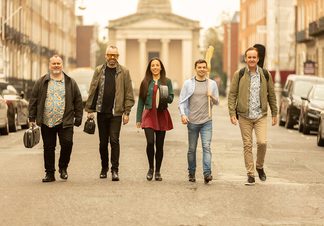Five people walking up a street, cars are parked on either side of the road.