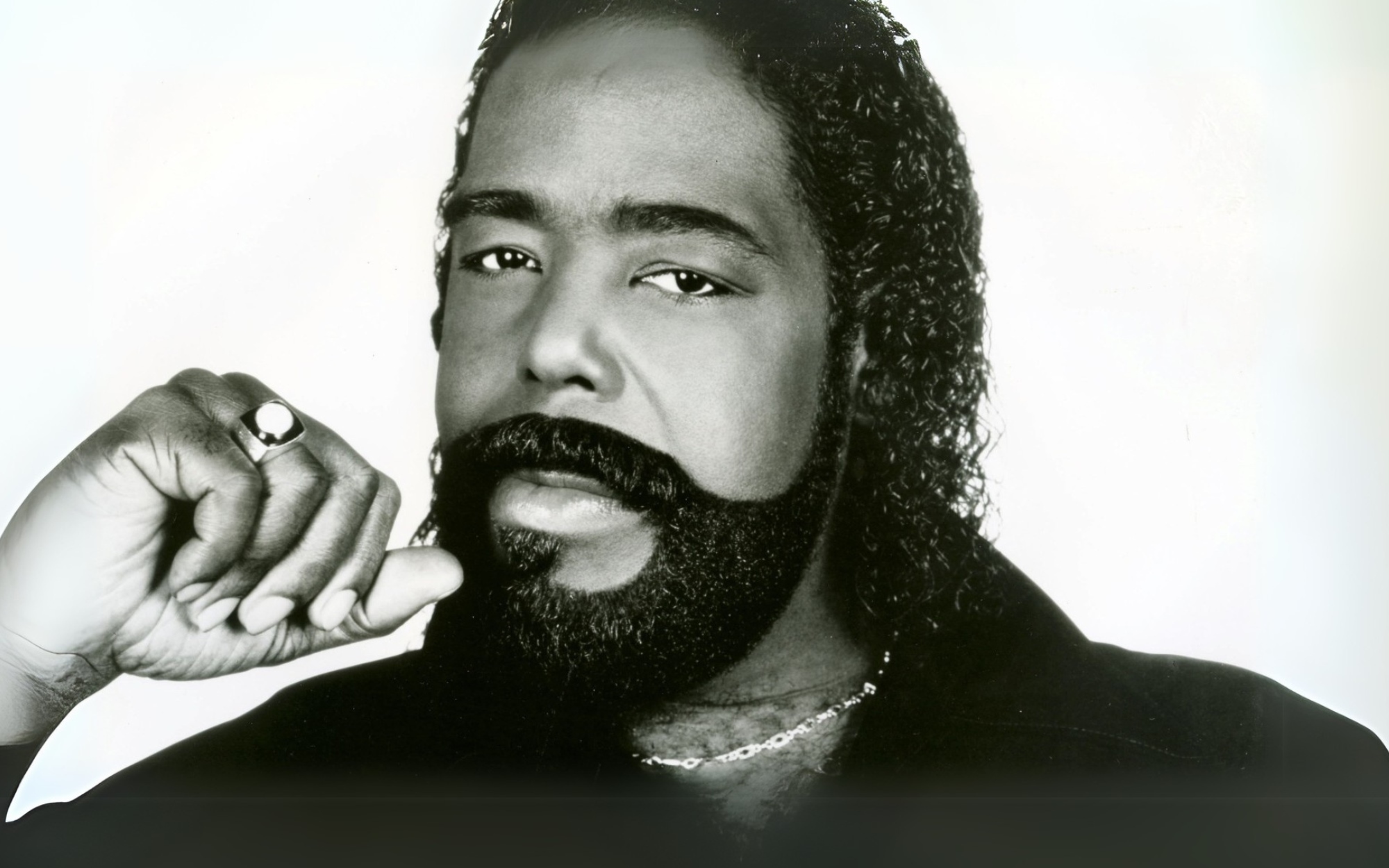 Black and white image of a man with a beard, wearing a black shirt and necklace.