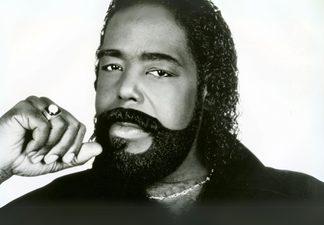 Black and white image of a man with a beard, wearing a black shirt and necklace.
