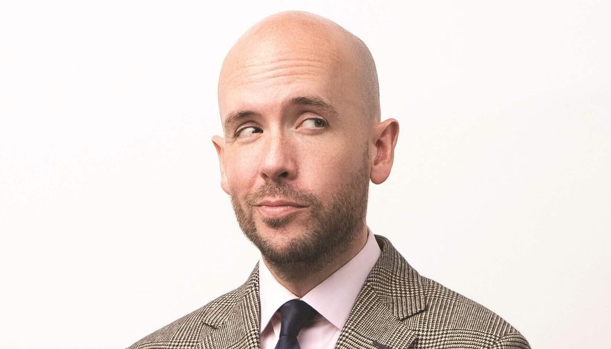 Headshot of Tom wearing a tweed suit, white shirt and navy blue tie.