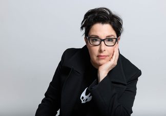 A person with short dark hair and black glasses poses against a light grey background, resting their face on one hand.