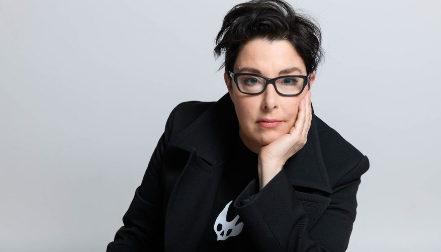 A person with short dark hair and black glasses poses against a light grey background, resting their face on one hand.